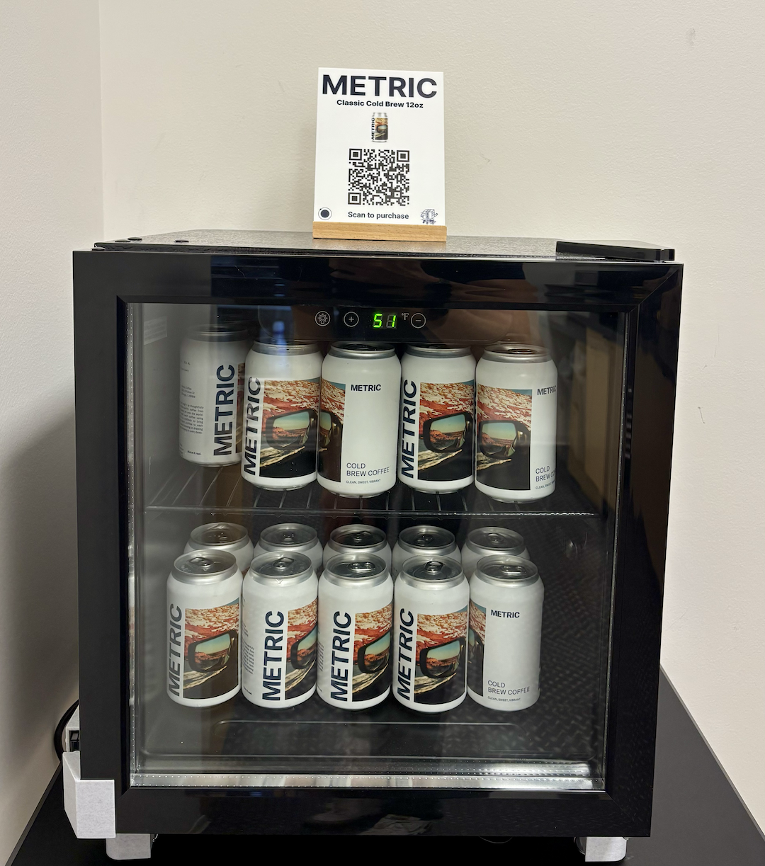 A glass-door beverage fridge stocked with cans of Metric cold brew, with a "Scan to purchase" QR code sign on top